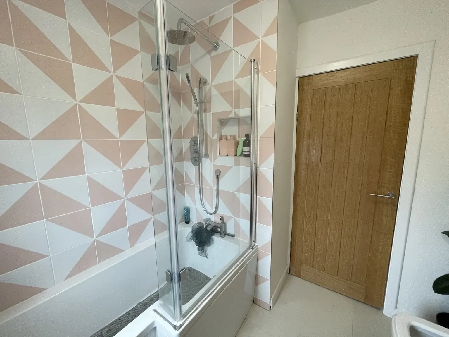 Bath and glass shower screen with pink-and-white geometric wall tiles and a recessed shower niche.