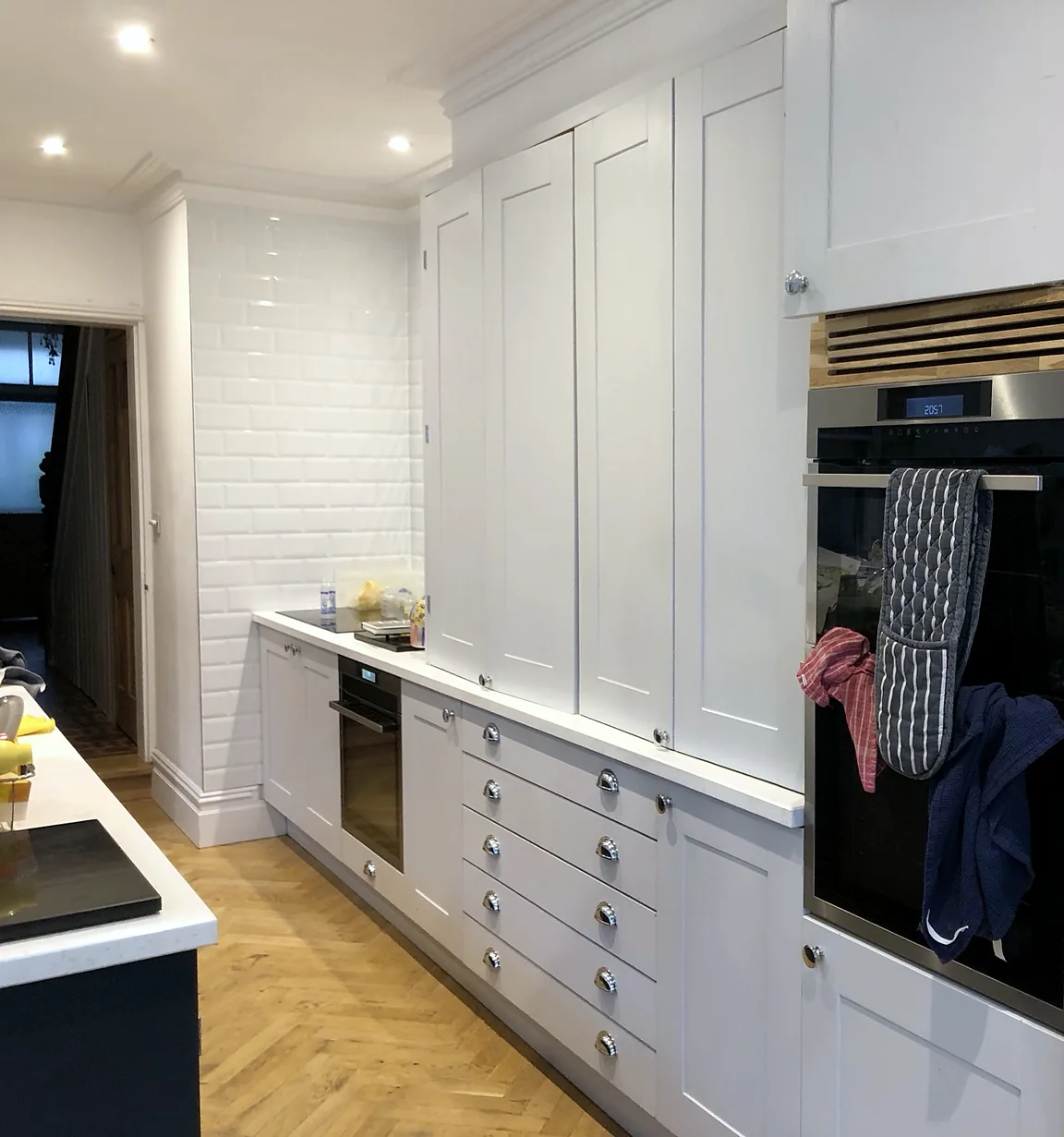 Tall pale shaker cabinets with a cornice above a built-in oven, beside a metro-tiled wall and oak herringbone floor.