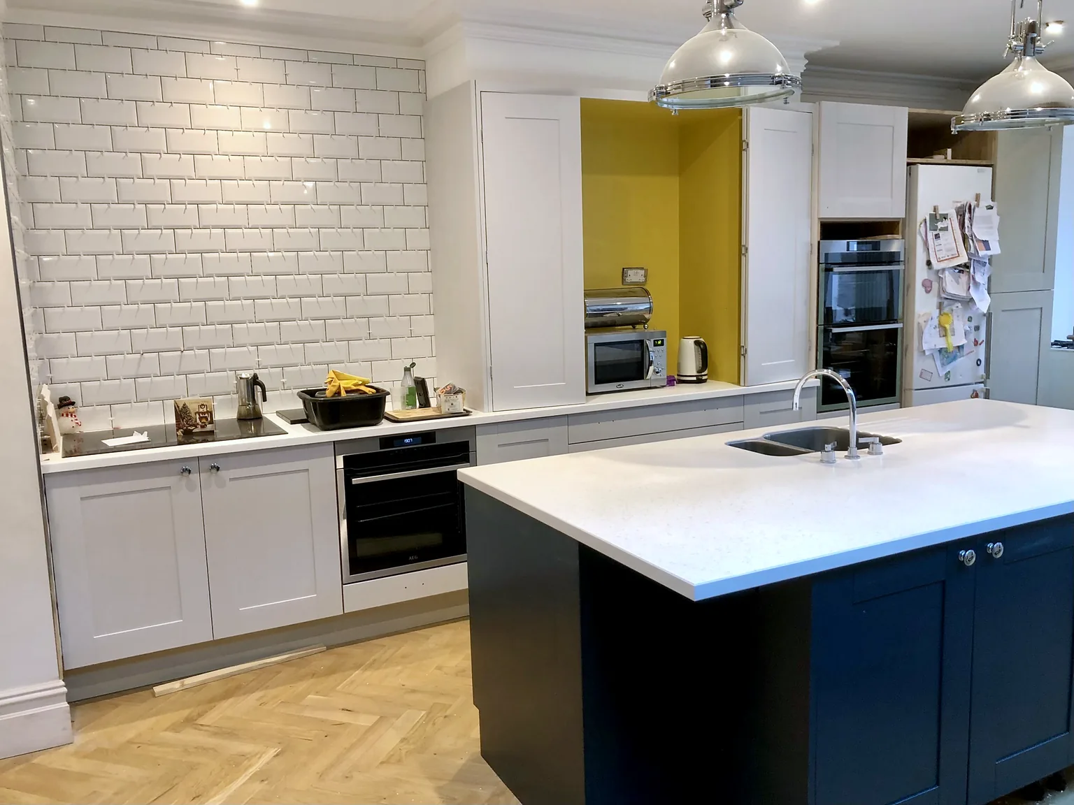 Kitchen with a metro-tiled wall, pale shaker cabinetry, a yellow-lined cupboard and a dark island with white worktop above herringbone flooring.